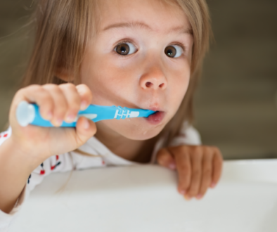 A little girl brushing her teeth over the sink with a surprised expression.